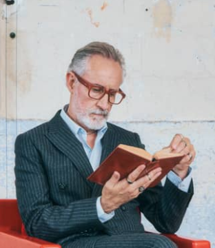 Man reading a book wearing red glasses in a chair with a neutral background