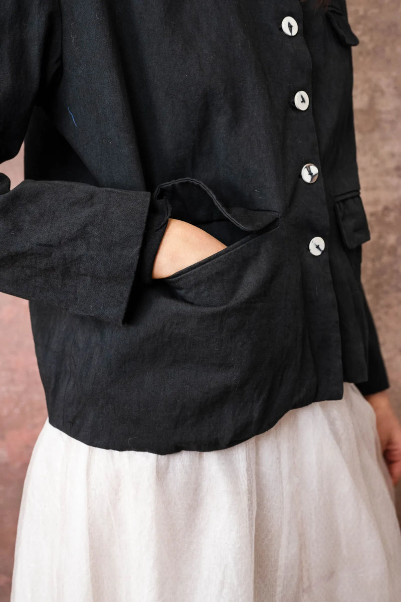 Person wearing a black jacket with white buttons and white pants against a brown background