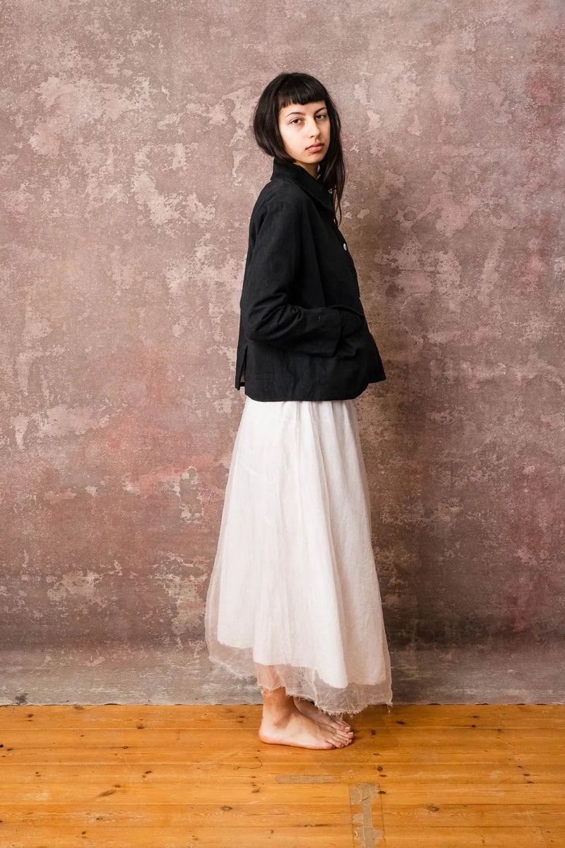 Woman wearing a black jacket and white skirt standing against a textured wall.
