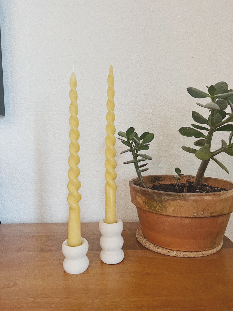 Two twisted candles in white holders on a wooden surface with a plant in the background.