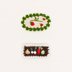 Two crocheted hair clips on a white background, one with a green border and red flowers, the other with a brown border and vegetables.
