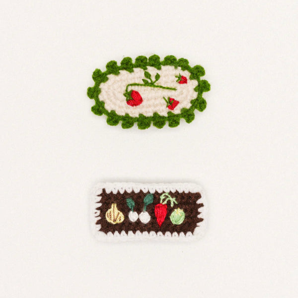 Two crocheted hair clips on a white background, one with a green border and red flowers, the other with a brown border and vegetables.