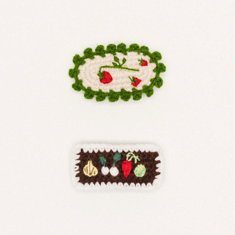 Two crocheted hair clips on a white background, one with a green border and red flowers, the other with a brown border and vegetables.