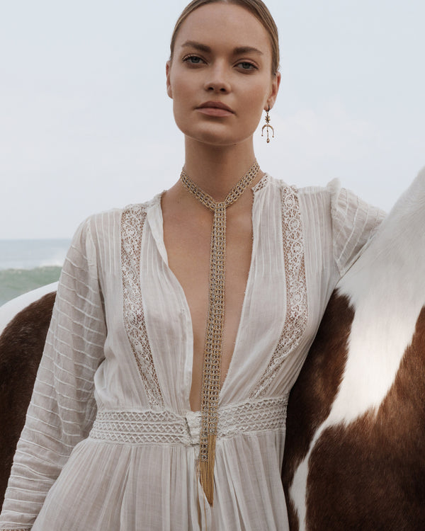 Woman in a white dress with intricate detailing and a gold wrap necklace standing next to a horse.